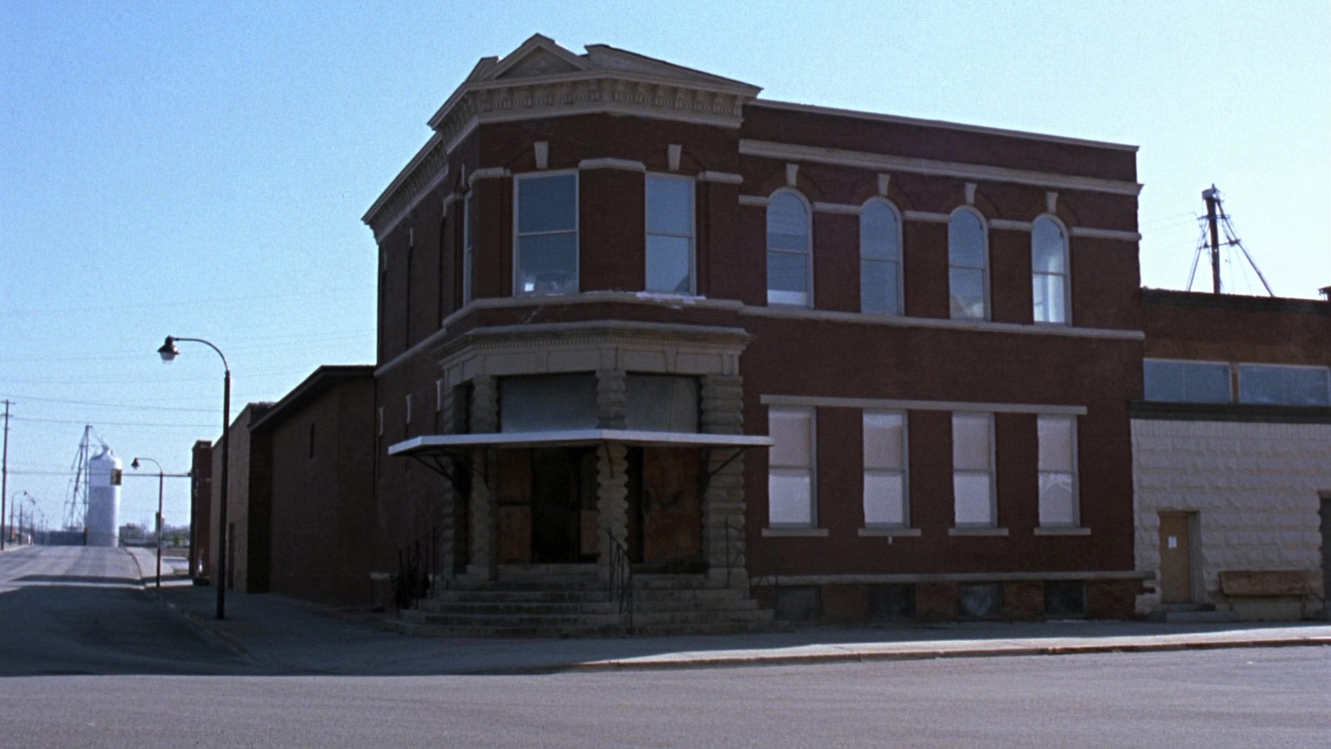 A small two-storey brick community bank on the main street of a rural Missouri town in morning light, with tall arched windows, stone lintels and a corner entrance with worn stone steps.
